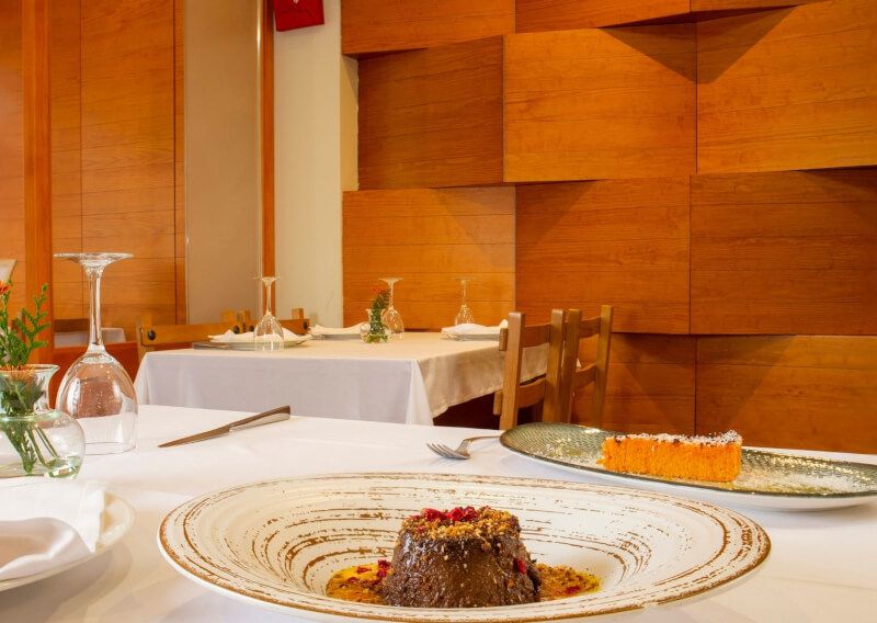 Fotografía de una mesa con dos postres en primer plano, otra mesa al fondo y la pared de madera.