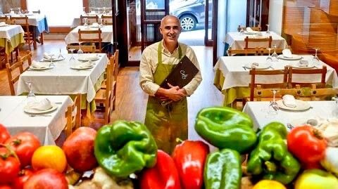 Fotografía del restaurante con vegetales en primer plano, mesas y dueño en segudo y detrás la puerta de entrada